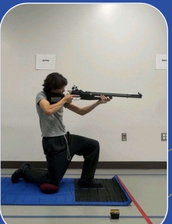 Kingwood NJROTC cadet firing an air rifle from a kneeling position on a blue mat
